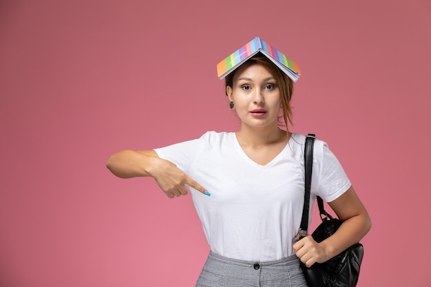 Vista frontal joven estudiante en camiseta blanca y pantalón gris con cuaderno en la cabeza sobre fondo rosa lecciones de estudiantes universitarios