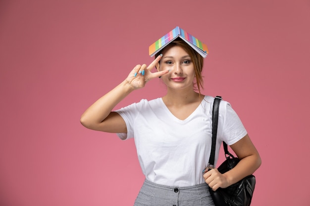 Vista frontal joven estudiante en camiseta blanca y pantalón gris con cuaderno en la cabeza sobre fondo rosa lecciones de estudiantes universitarios