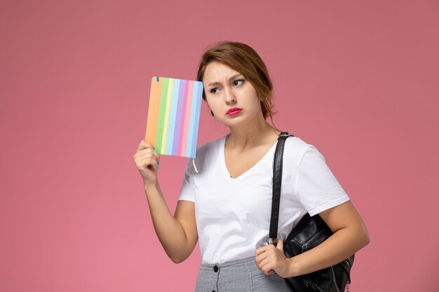 Vista frontal joven estudiante en camiseta blanca y pantalón gris con cuaderno en sus manos pensando en el fondo rosa lecciones universitarias