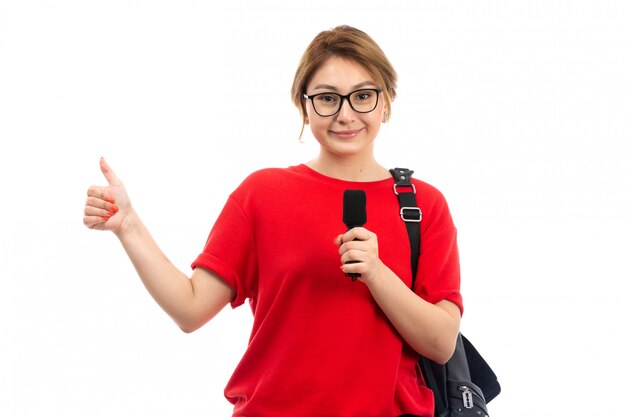 Una vista frontal joven estudiante en camiseta roja con bolsa negra con micrófono sonriendo en el blanco