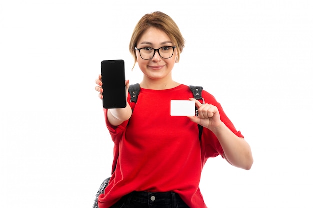 Una vista frontal joven estudiante en camiseta roja con bolsa negra con smartphone negro y tarjeta blanca sonriendo en el blanco