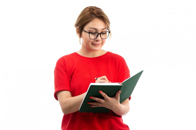 Una vista frontal joven estudiante en camiseta roja con cuaderno escribiendo notas sobre el blanco