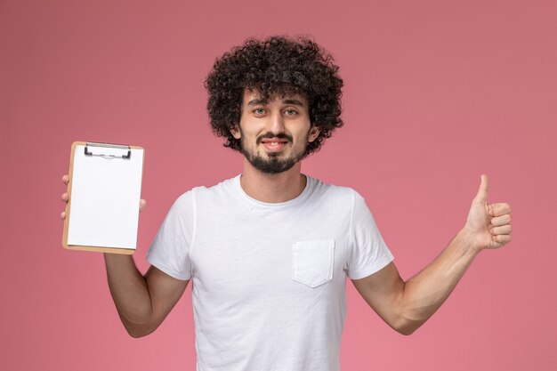 Vista frontal joven estudiante dando pulgares al cuaderno blanco sobre fondo rosa aislado