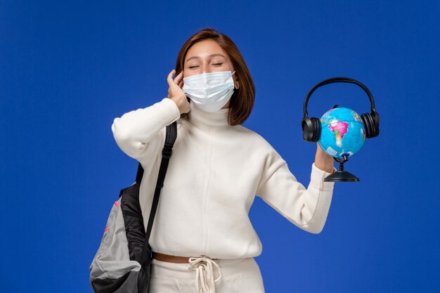 Vista frontal joven estudiante en jersey blanco con máscara y mochila sosteniendo globo con auriculares en la pared azul