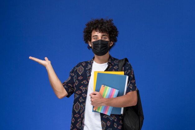 Vista frontal joven estudiante con máscara negra con mochila con archivos y cuaderno sobre fondo azul.