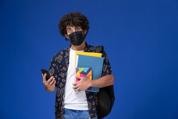 Vista frontal joven estudiante con máscara negra con mochila con archivos y teléfono sobre fondo azul.