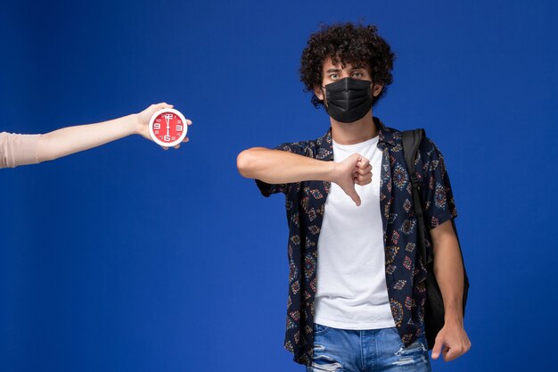 Vista frontal joven estudiante con máscara negra con mochila mostrando signo diferente sobre fondo azul claro.