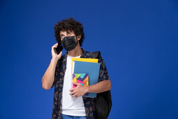 Vista frontal joven estudiante con máscara negra con mochila sosteniendo archivos y hablando por teléfono sobre fondo azul.