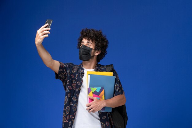 Vista frontal joven estudiante con máscara negra con mochila sosteniendo archivos y tomando selfie sobre fondo azul.