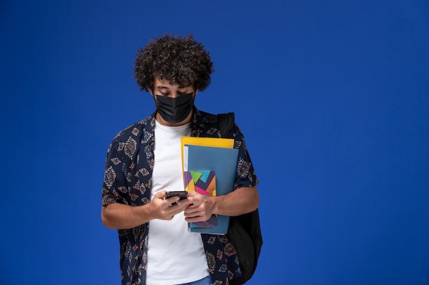 Foto gratuita vista frontal joven estudiante con máscara negra con mochila sosteniendo archivos y usando su teléfono en el fondo azul.