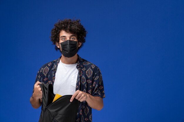 Foto gratuita vista frontal joven estudiante con máscara negra y sosteniendo una mochila negra sobre fondo azul claro.