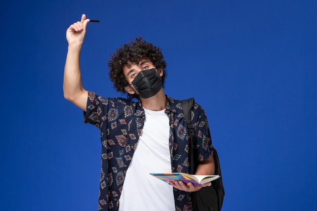 Vista frontal joven estudiante vistiendo máscara negra con mochila con cuaderno sobre fondo azul.