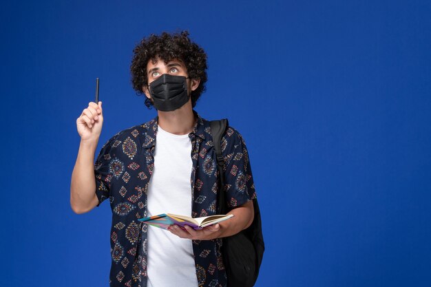 Vista frontal joven estudiante vistiendo máscara negra con mochila con cuaderno sobre fondo azul.