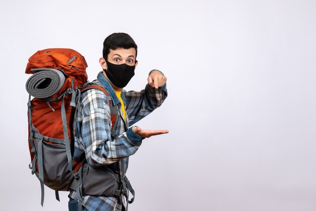 Vista frontal joven excursionista con mochila y máscara negra de pie sobre fondo blanco.