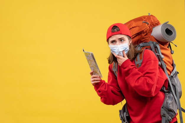 Vista frontal joven excursionista con mochila y máscara sosteniendo el mapa poniendo la mano en la barbilla