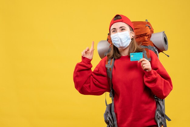 Vista frontal joven excursionista con mochila y máscara con tarjeta de crédito apuntando al techo