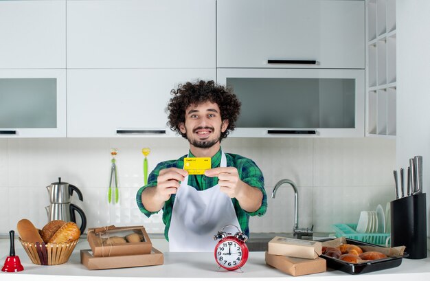 Vista frontal del joven feliz de pie detrás de la mesa varios pasteles y mostrando la tarjeta bancaria en la cocina blanca