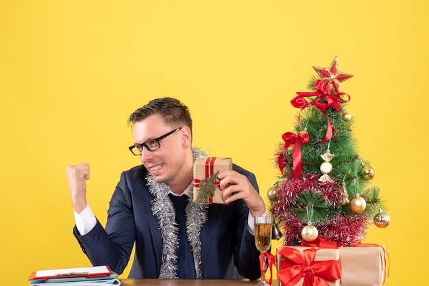 Vista frontal del joven feliz sentado en la mesa cerca del árbol de Navidad y presenta en amarillo