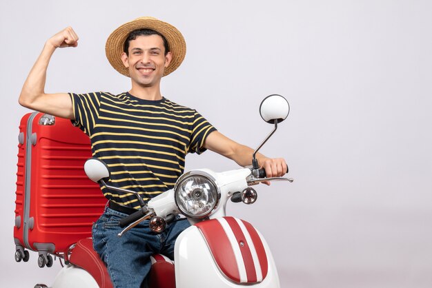 Vista frontal del joven feliz con sombrero de paja en ciclomotor mostrando el músculo del brazo