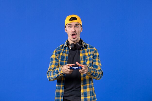 Vista frontal joven con gamepad y auriculares jugando videojuegos en la pared azul