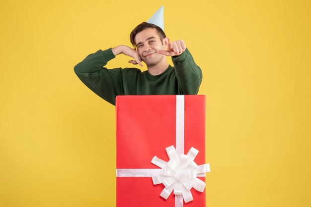 Vista frontal joven con gorra de fiesta apuntando a la cámara de pie detrás de una gran caja de regalo sobre fondo amarillo