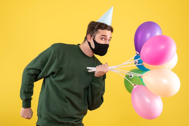 Vista frontal joven con gorra de fiesta azul y globos de colores de pie en amarillo