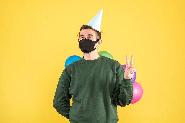 Vista frontal joven con gorra de fiesta escondiendo globos detrás de su espalda de pie sobre fondo amarillo