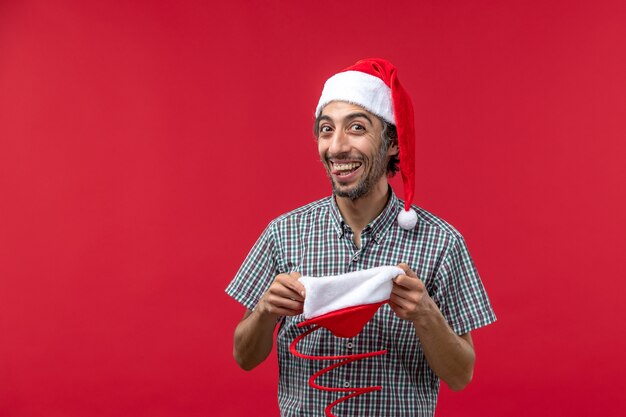 Vista frontal del joven con gorra roja de juguete de navidad en la pared roja