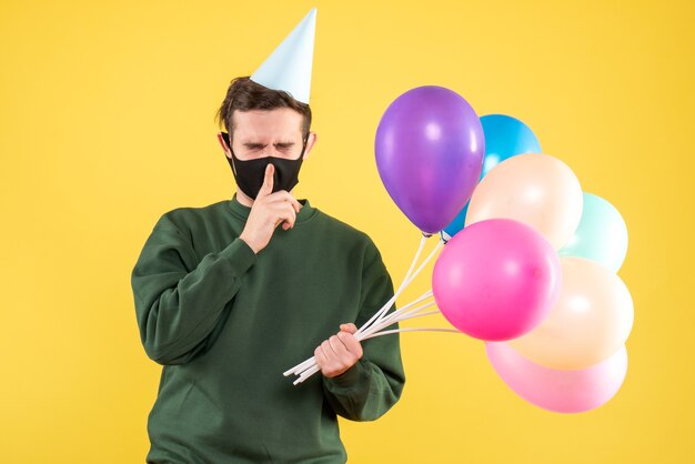 Vista frontal joven con gorro de fiesta y globos de colores haciendo cartel de shh de pie en amarillo