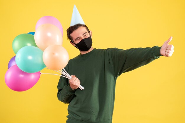 Vista frontal joven con gorro de fiesta y globos de colores haciendo pulgar arriba cartel de pie en amarillo