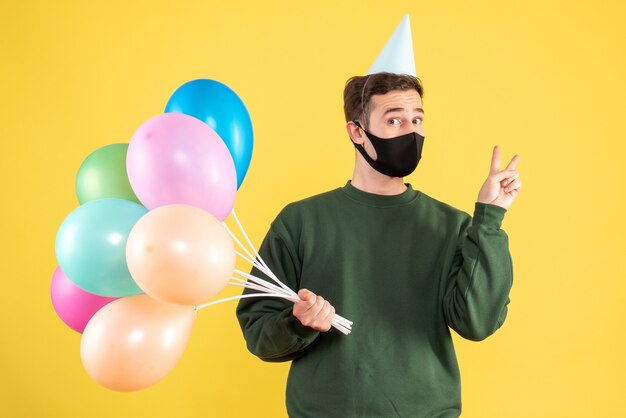 Vista frontal joven con gorro de fiesta y globos de colores haciendo el signo de la victoria de pie en amarillo