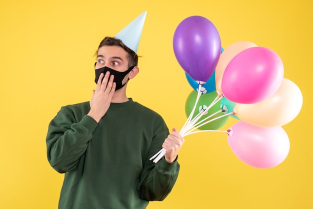 Vista frontal joven con gorro de fiesta y globos de colores de pie en amarillo