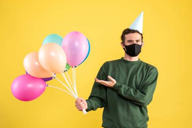 Vista frontal joven con gorro de fiesta y globos de colores de pie sobre fondo amarillo