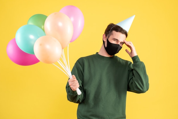 Vista frontal joven con gorro de fiesta y globos de colores sosteniendo la cabeza de pie en amarillo