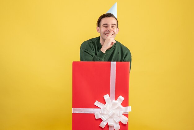 Vista frontal joven con gorro de fiesta haciendo signo shh de pie detrás de caja de regalo grande sobre fondo amarillo