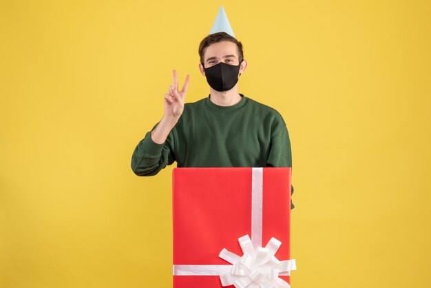 Vista frontal joven con gorro de fiesta haciendo el signo de la victoria de pie detrás de una gran caja de regalo sobre fondo amarillo