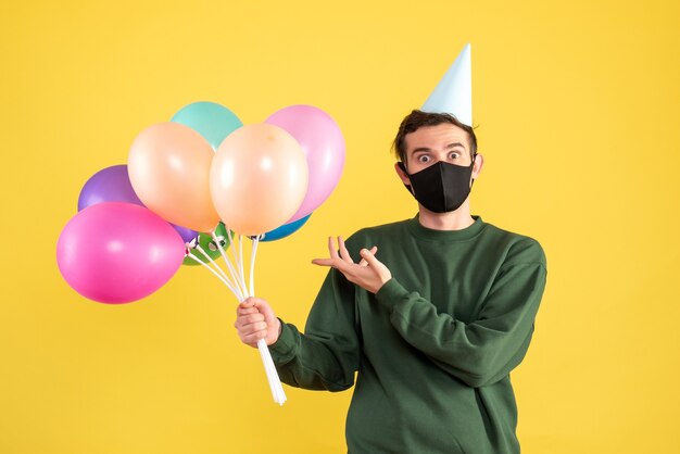 Vista frontal joven con gorro de fiesta y máscara negra apuntando a globos en amarillo