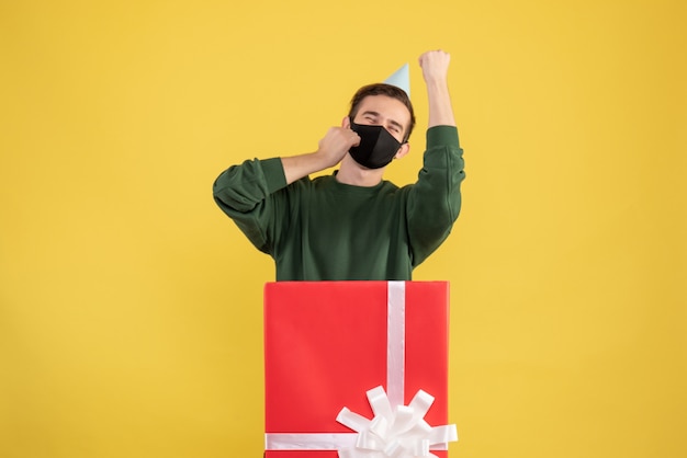 Vista frontal joven con gorro de fiesta y máscara negra de pie detrás de caja de regalo grande en amarillo