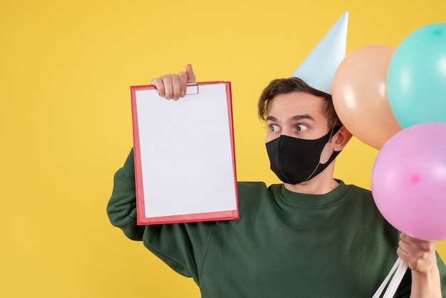 Vista frontal joven con gorro de fiesta y máscara negra con portapapeles y globos en amarillo