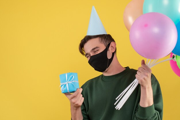 Vista frontal joven con gorro de fiesta y máscara negra con regalo y globos en amarillo