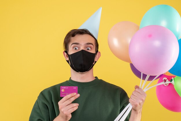 Vista frontal joven con gorro de fiesta y máscara negra con tarjeta y globos en amarillo