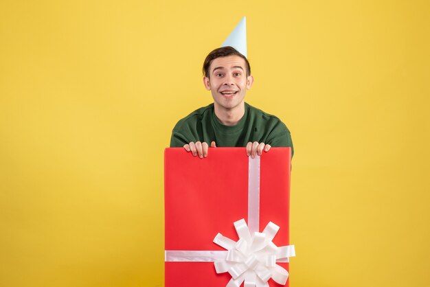 Vista frontal joven con gorro de fiesta de pie detrás de una gran caja de regalo sobre fondo amarillo