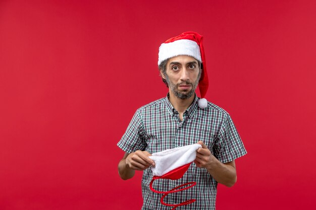 Vista frontal del joven con gorro de juguete de navidad en la pared roja