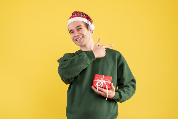 Vista frontal joven con gorro de Papá Noel apuntando a algo sobre fondo amarillo