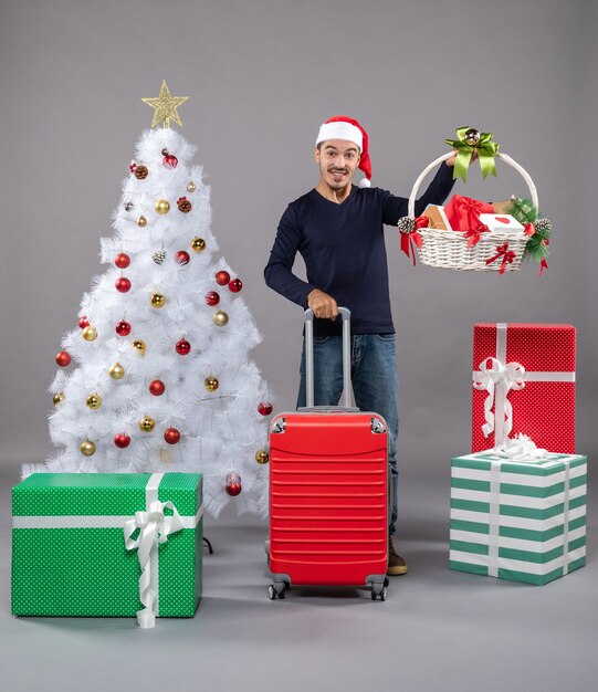 Vista frontal joven con gorro de Papá Noel con canasta de regalo y bolsa de viaje roja en aislado