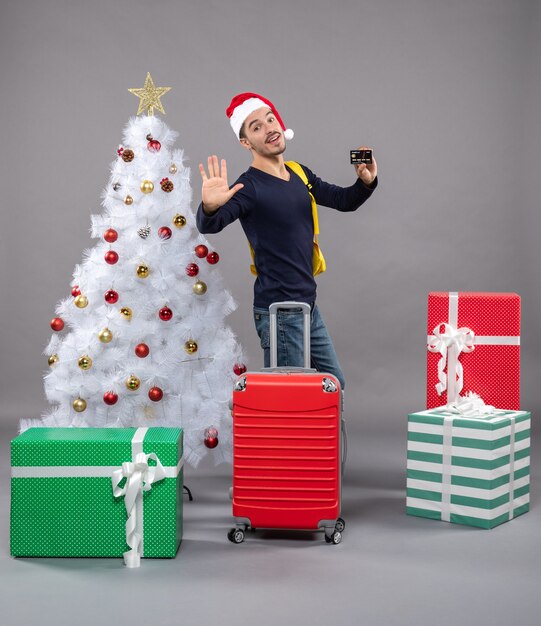 Vista frontal joven con gorro de Papá Noel y maleta roja cerca del árbol de Navidad blanco en gris aislado