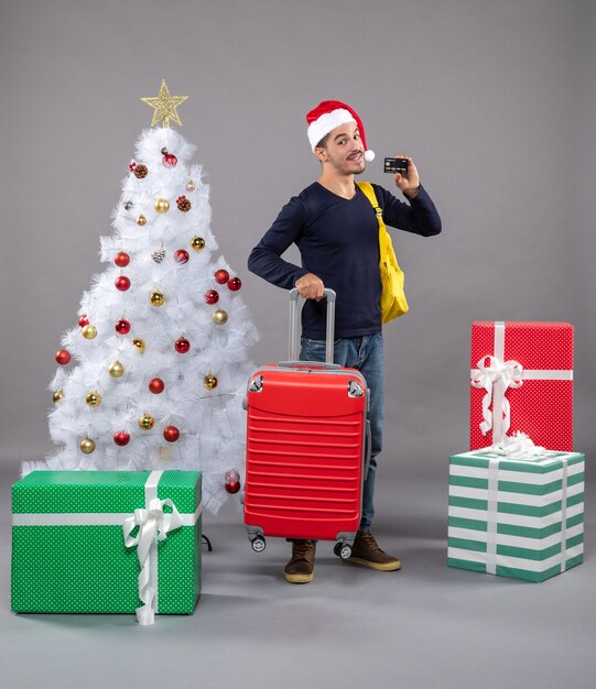 Vista frontal joven con gorro de Papá Noel con maleta roja mostrando una tarjeta en aislado