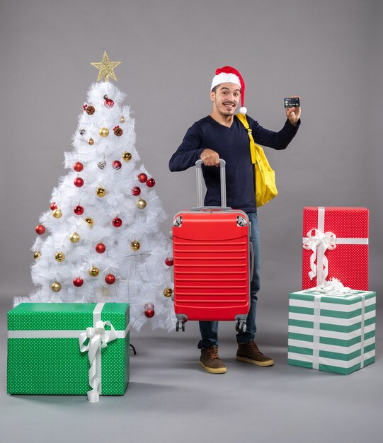 Vista frontal joven con gorro de Papá Noel con maleta roja mostrando una tarjeta alrededor de coloridos regalos en gris aislado