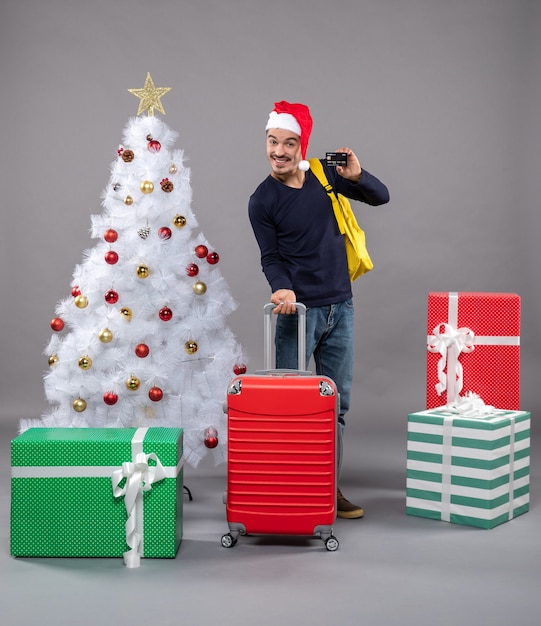 Vista frontal joven con gorro de Papá Noel con maleta roja mostrando una tarjeta en gris aislado