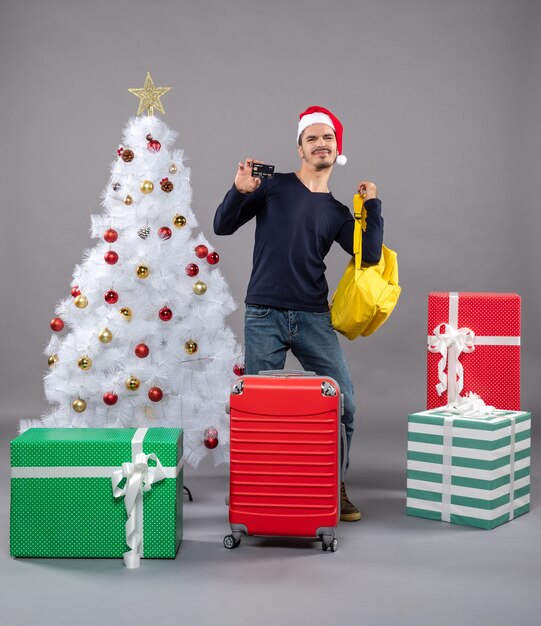 Vista frontal joven con gorro de Papá Noel y maleta roja tomando su mochila amarilla en gris aislado